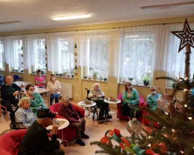 Grupa seniorów i pracowników siedzi w fotelach i ogląda film przygotowany przez dzieci z przedszkola Lokomotywa z życzeniami świątecznymi i kolędami