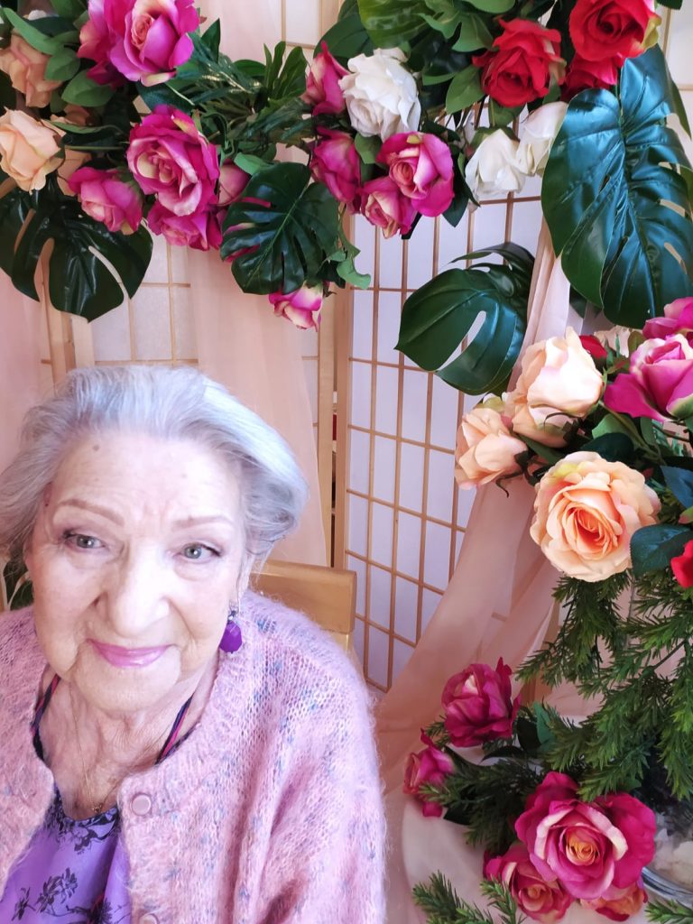 Na krześle siedzi uśmiechnięta seniorka. Ma na sobie różowy sweterek i kolorystycznie dobrany makijaż. W tle widać dekorację z zielonych gałązek, białych, czerwonych, różowych i kremowych róż