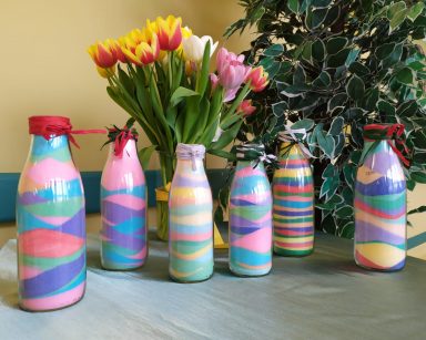 Na blacie stoją szklane przezroczyste butelki. W butelkach jest kolorowa sól: zielona, żółta, niebieska, różowa, pomarańczowa. Sól ułożona jest warstwami. W tle widać wazon z żółtymi, białymi i różowymi tulipanami