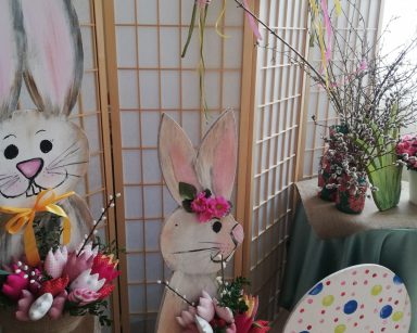 Dekoracja świąteczna: dwa drewniane zające trzymają bukiety kolorowych kwiatów. Przy nich kolorowa drewniana pisanka. Obok stolik z różowymi kwiatami, baziami i gałązkami brzozowymi w wazonach