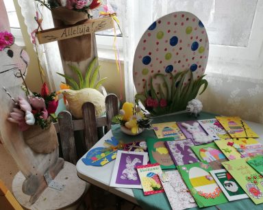 Na stole leżą rozłożone kartki świąteczne. Są ozdobione obrazkami wielkanocnych zajączków, baranków, pisanek, kwiatów i bazi. Na blacie widać jeszcze białego baranka i żółty stroik. Obok stołu stoją drewniane dekoracje: zając z bukietem materiałowych kwiatów, pomalowana na żółto kura, duża biała pisanka w niebieskie zielone i różowe kropki, tabliczka z napisem Alleluja!