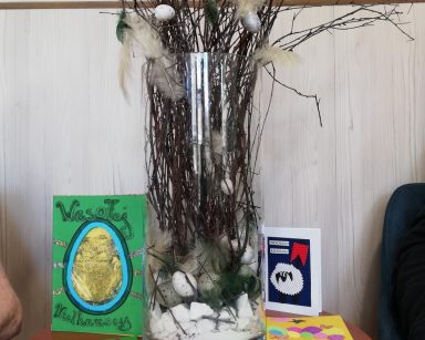 Na środku stołu stoi szklany przezroczysty wazon z gałązkami leszczyny. Gałązki są udekorowane kremowymi piórkami, białymi i srebrnymi pisankami. Obok na blacie leżą kolorowe wielkanocne kartki z życzeniami