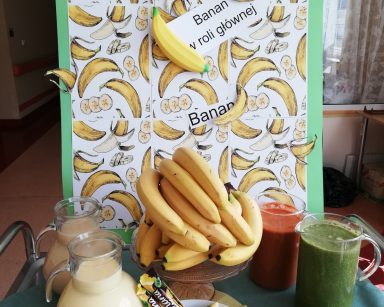Plakat, na białym tle żółte banany i napis: Banan w roli głównej. Na stole soki, bananowe chrupki, serki i batony.