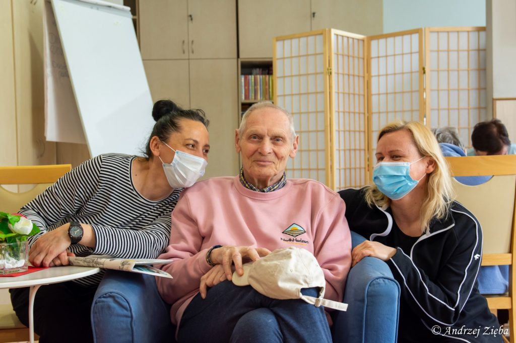 Jasny pokój. Terapeutka Magdalena Poraj-Górska, senior, neurologopedka Anna Szmaja-Wysocka siedzą obok siebie uśmiechnięci.