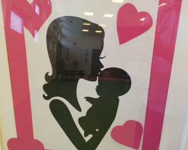 Plakat. Na białym tle kobieca postać całująca noworodka. Wokół różowa ramka i różowe serca.