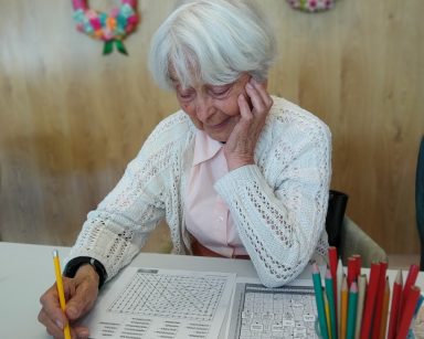 Seniorka siedzi przy stole i rozwiązuje wykreślankę. Obok gazeta z krzyżówką i kolorowe ołówki.