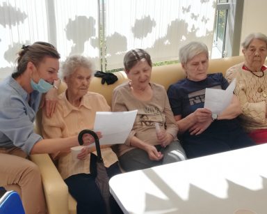 Psycholożka Maria Skubich-Wiczling i cztery seniorki siedzą na kanapie. Dwie trzymają w dłoniach kartki z tekstem piosenki.