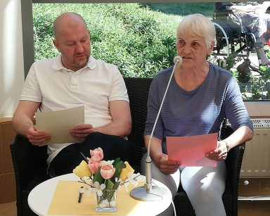 Seniorka odczytuje życzenia. Obok kierownik Arkadiusz Wanat. Przed nimi stolik z bukietem kolorowych kwiatów.