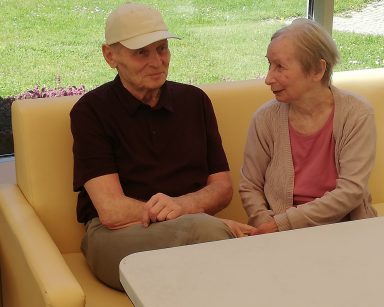 Dwoje uśmiechniętych seniorów siedzi obok siebie na kanapie.