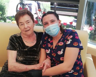 Seniorka i fizjoterapeutka Martyna Józefczyk siedzą na kanapie. Trzymają się za ręce i uśmiechają.