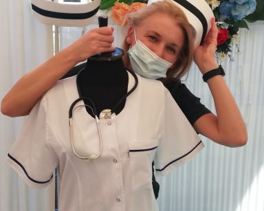 Kierowniczka Ilona Gajewska. Jedną ręką przytrzymuje czepek pielęgniarski drugą chwyta stojak z białym fartuchem.
