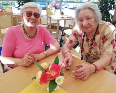 Dwie roześmiane seniorki przy stole. Na blacie wazon z bukietem kolorowych kwiatów.
