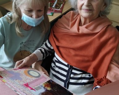 Terapeutka Ewa Rodziewicz i seniorka oglądają kolorowy album. W albumie zdjęcia seniorki.