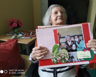 Seniorka prezentuje album ze swoim zdjęciem. Jest ozdobione kolorowym papierem, wstążką i wyciętymi obrazkami kwiatów.