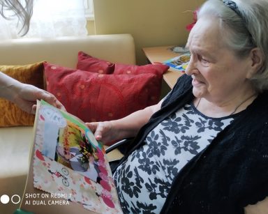 Seniorka ogląda album ze swoim zdjęciem i uśmiecha się. Zdjęcie jest udekorowane kolorowymi, papierowymi ozdobami.