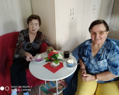Dwie roześmiane seniorki siedzą przy stoliku. Na blacie szklanki z kawą, babeczki, wazon z kwiatami.
