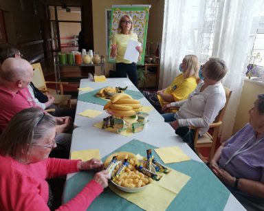 Kierowniczka Ilona Gajewska prowadzi spotkanie. Seniorzy i pracownicy siedzą przy stole. Na blacie bananowe przekąski.