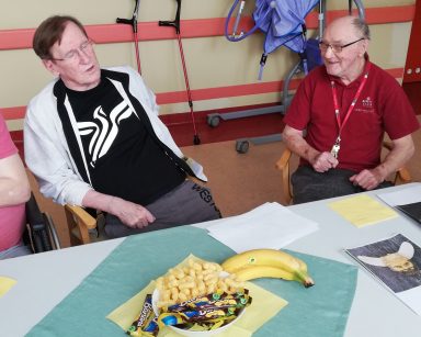 Dwóch seniorów rozmawia siedząc przy stole. Na blacie banany, bananowe chrupki i batony.