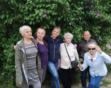 Seniorzy i terapeutka Beata Gadomska stoją objęci. Za nimi wysoki krzew z fioletowym bzem.