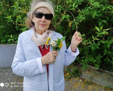 Seniorka stoi na tle zielonych krzewów. W dłoni trzyma bukiecik polnych kwiatów.