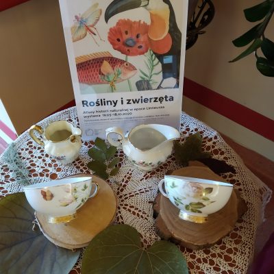 Na stoliku plakat reklamujący wystawę. Obok dekoracja: filiżanki i dzbanuszki na mleko z kwiatowymi wzorami, zielone liście.