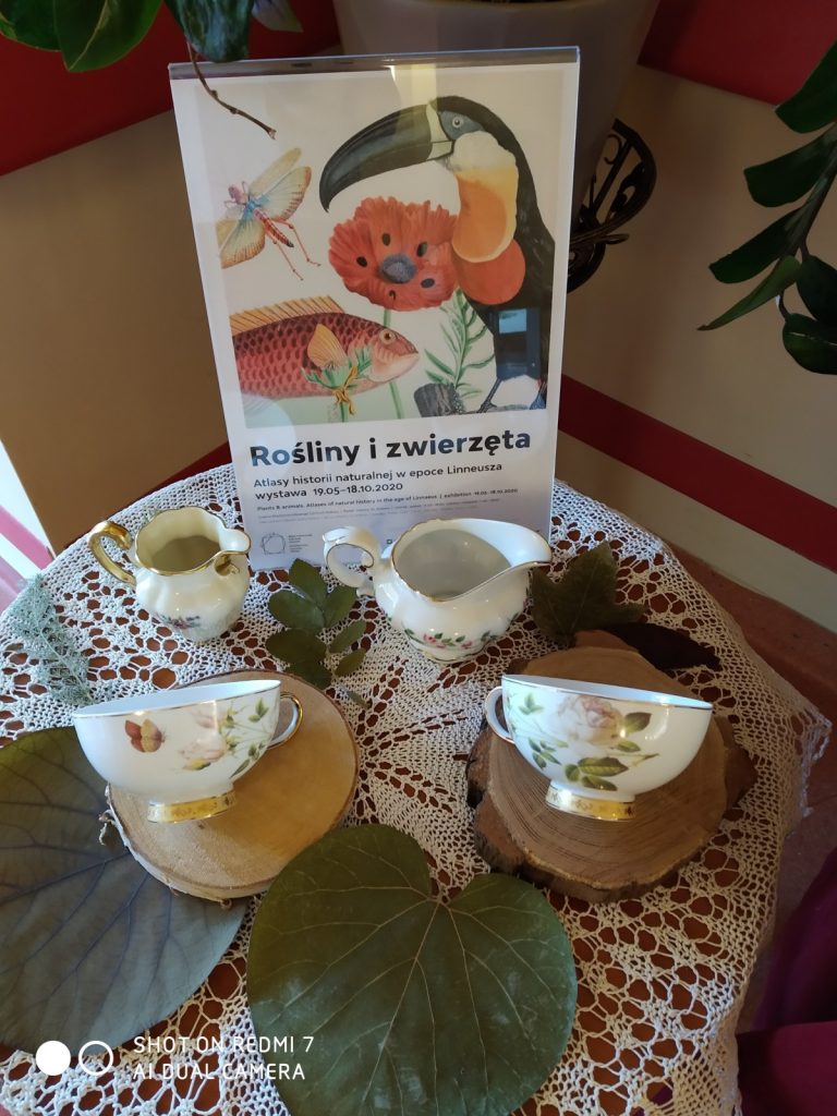 Na stoliku plakat reklamujący wystawę. Obok dekoracja: filiżanki i dzbanuszki na mleko z kwiatowymi wzorami, zielone liście.