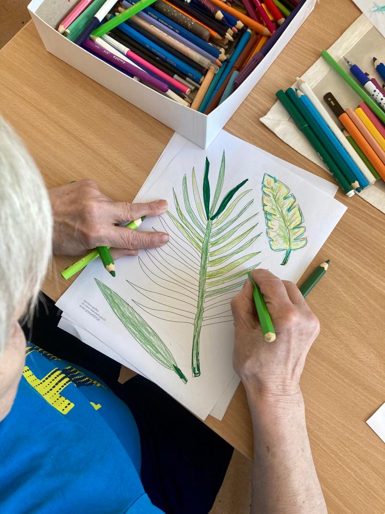 Seniorka pochyla się nad stołem, w dłoni trzyma zielone kredki. Na blacie rozpoczęty rysunek, kolorowe kredki.