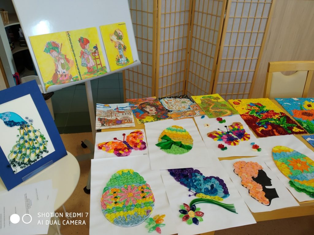 Na stoliku, stole i stelażu kolorowe obrazki. Obrazki wykonane są z bibuły, pasków kolorowego papieru, farb i kredek.