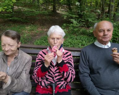 Na ławce w lesie siedzi troje seniorów. Jedzą lody.