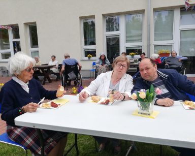Seniorzy siedzą przy stolikach na patio przed DPS. Jedzą kiełbaski z grilla.