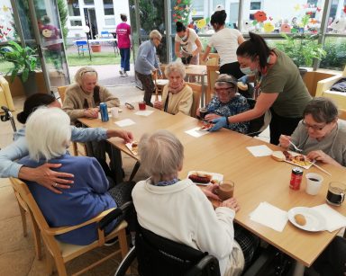 Ogród zimowy. Przy stołach seniorzy, wolontariusze i pracownicy, jedzą kiełbaski z grilla, rozmawiają.