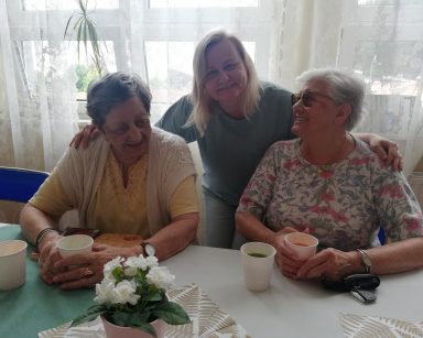Przy stole dwie seniorki i neurologopedka Anna Szmaja-Wysocka. Śmieją się, w rękach mają kubki z sokami. Za nimi duże okna.
