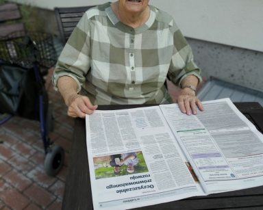 Na patio przy stoliku seniorka. Przed nią rozłożona gazeta.