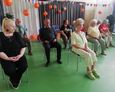 Sala gimnastyczna. Dekoracja z balonów i kolorowych chorągiewek. Seniorzy siedzą na krzesłach.