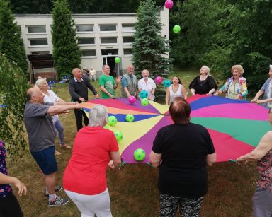 Trawnik. Seniorzy i pracownicy podrzucają piłki wspólnie trzymając kolorową chustę.