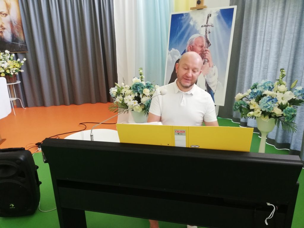 Kierownik Arkadiusz Wanat gra na pianinie. W tle portret papieża Jana Pawła II i dekoracje z kwiatów.