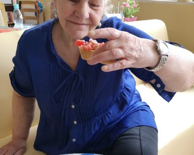 Seniorka siedzi na kanapie. W dłoni trzyma kanapkę z pokrojonymi pomidorami, przed nią na tacce ser mozzarella.