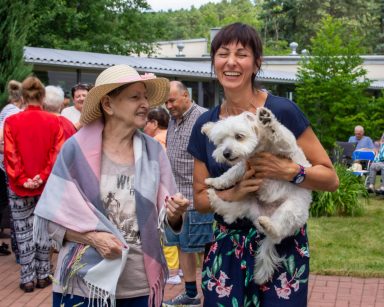 Terapeutka Ania Rzepczyńska śmieje się z psem Mitsu na rękach. Obok uśmiechnięta seniorka. Za nimi seniorzy i pracownicy.