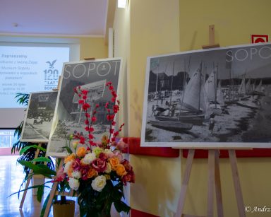 Na stelażach trzy czarno-białe plakaty ze zdjęciami miasta, plaży i napisem Sopot. Obok wazon z kwiatami. W tle ekran projektora.