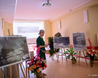 Edukatorka Iwona Zając prowadzi spotkanie. Stoi między plakatami ze zdjęciami Sopotu i jego okolic. Za nią ekran projektora.