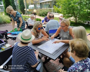Na patio w ogrodzie dyrektorka Agnieszka Cysewska, seniorzy i pracownicy. Grają przy stolikach w gry planszowe.