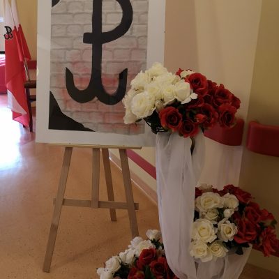 Dekoracja. Plakat z symbolem Polski Walczącej. Obok bukiety białych i czerwonych róż ozdobione białym tiulem.