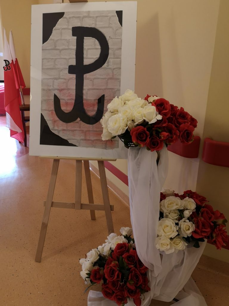 Dekoracja. Plakat z symbolem Polski Walczącej. Obok bukiety białych i czerwonych róż ozdobione białym tiulem.