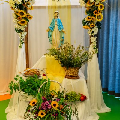 Pergola ozdobiona białymi i żółtymi kwiatami. Na podwyższeniu figurka Matki Boskiej. Poniżej kosze z darami do poświęcenia.