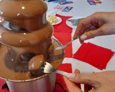 Na stole fontanna z płynną czekoladą. Widać dłonie pracownika, który macza w czekoladzie owoce nabite na widelce.