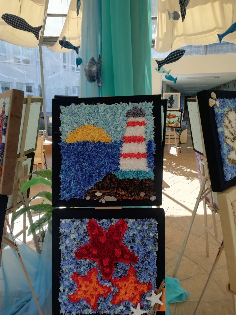 Ogród zimowy. Na sztalugach obrazy o tematyce morskiej wykonane z tkanin. Nad nimi dekoracja, kolorowe rybki z materiału.