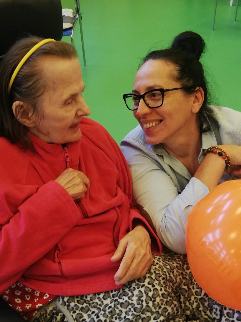 Na sali gimnastycznej obok siebie terapeutka Magdalena Poraj-Górska i seniorka. Uśmiechają się do siebie.