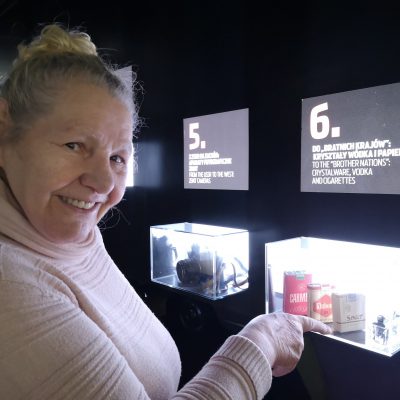 Uśmiechnięta seniorka wskazuje palcem wystawę w Muzeum Emigracji. Eksponaty to paczki papierosów, kryształy do wódki.