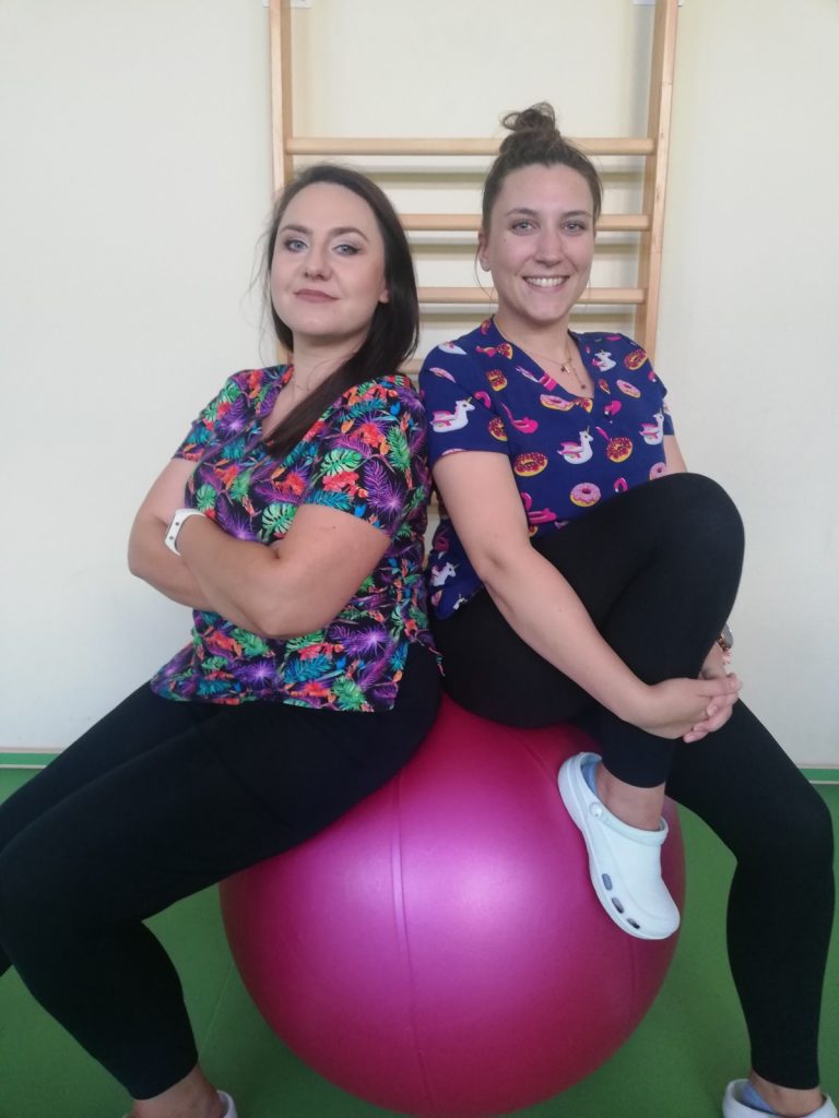 Sala gimnastyczna. Fizjoterapeutki Paulina Winczura i Martyna Józefczyk siedzą na dużej, różowej piłce. Uśmiechają się.