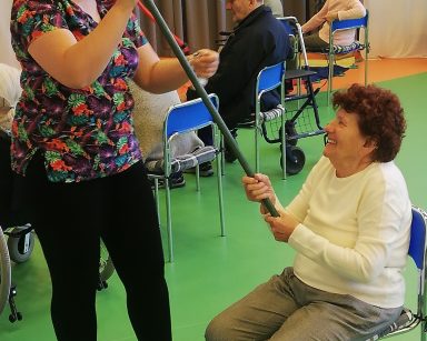 Fizjoterapeutka Paulina Winczura pomaga seniorce z wędką. W dalszej części sali gimnastycznej siedzą seniorzy.
